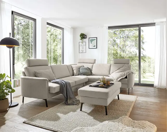Beiges Ecksofa mit Kissen und Hocker auf Teppich im Wohnzimmer, große Fensterfront, Stehlampe links und Sideboard rechts
